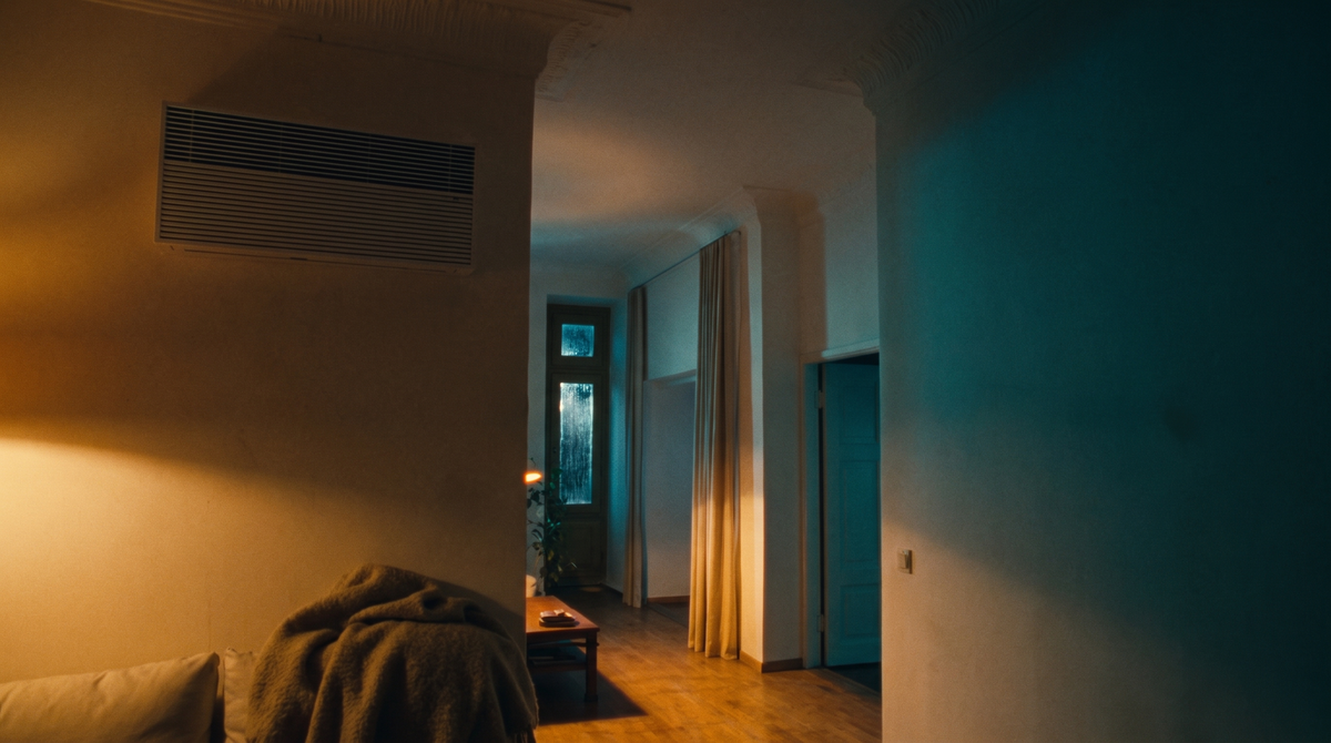 Dasselbe Wohnzimmer am Winterabend, warmes Lampenlicht, Regen am Fenster, Decke auf dem Sofa
